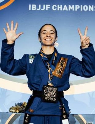 Mayssa Bastos no podio fazendo o sinal de sete com os dedos, representando o setimo titulo mundial