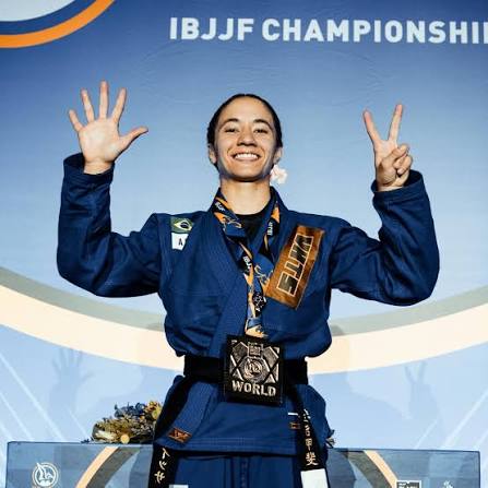 Mayssa Bastos no podio fazendo o sinal de sete com os dedos, representando o setimo titulo mundial